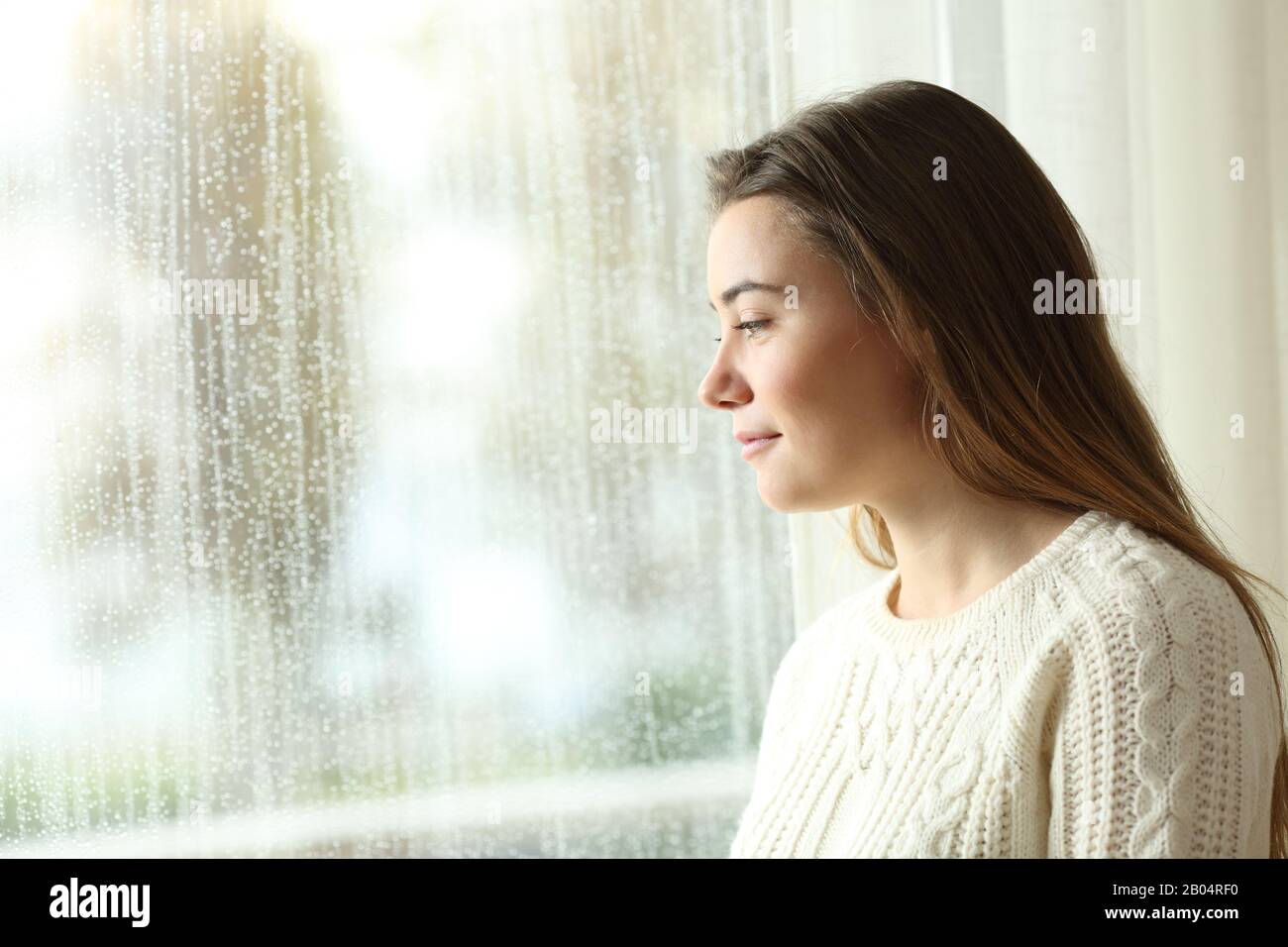 Side view portrait of a happy pensive girl looking the rain through a window at home Stock Photo