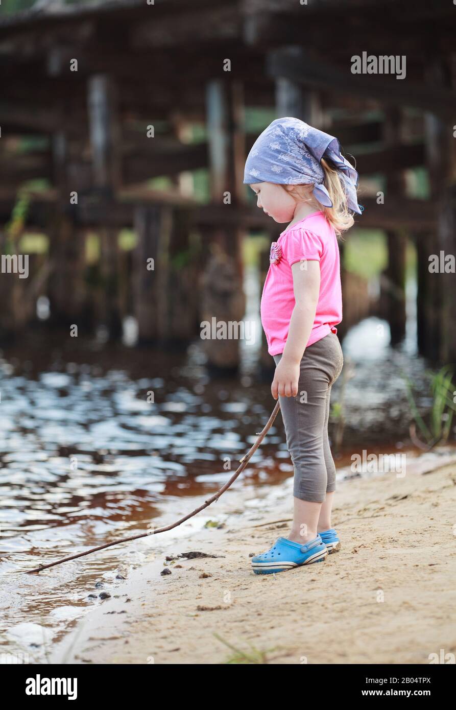 Little sad girl in a blue bandana stands on the bank of the river and draws  with a twig on the water Stock Photo - Alamy