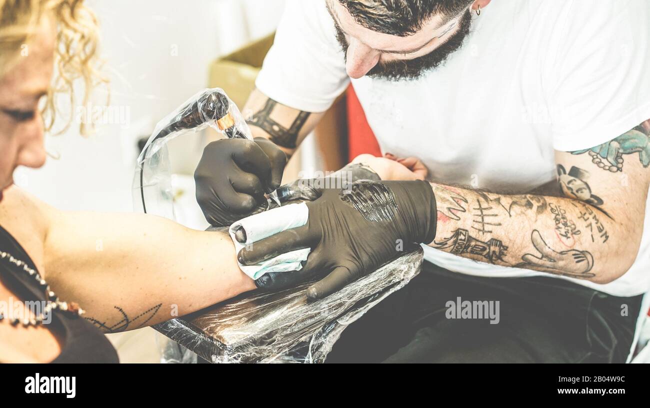 Bearded tattoo artist making tattoo inside ink studio on young blond woman - Tattooer at work - Skin trend fashion concept - Focus on man hand machine Stock Photo