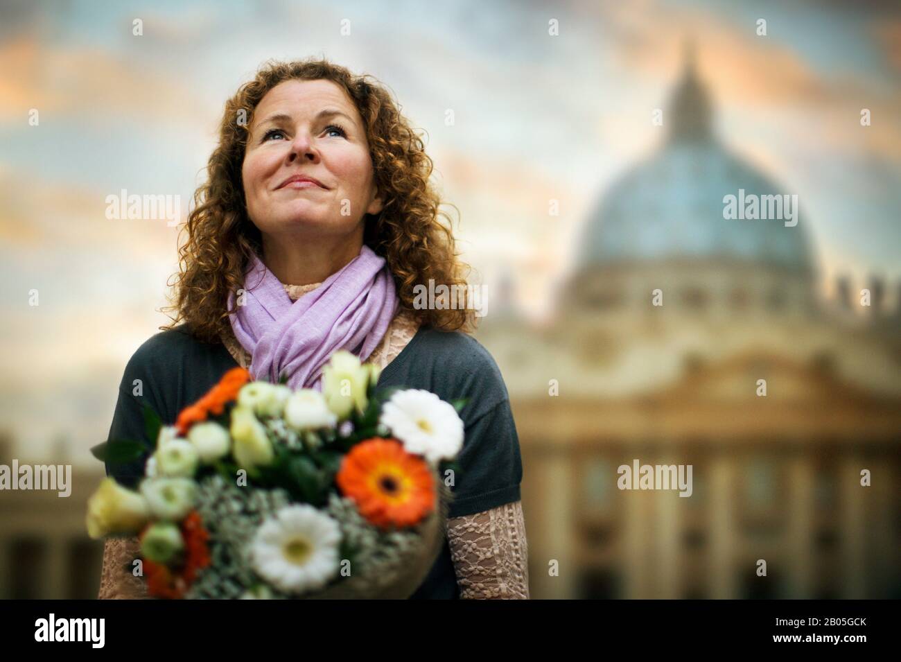 Smiling middle-aged woman holds a bouquet of flowers as she waits for someone outside the Sistine Chapel at sunset. Stock Photo