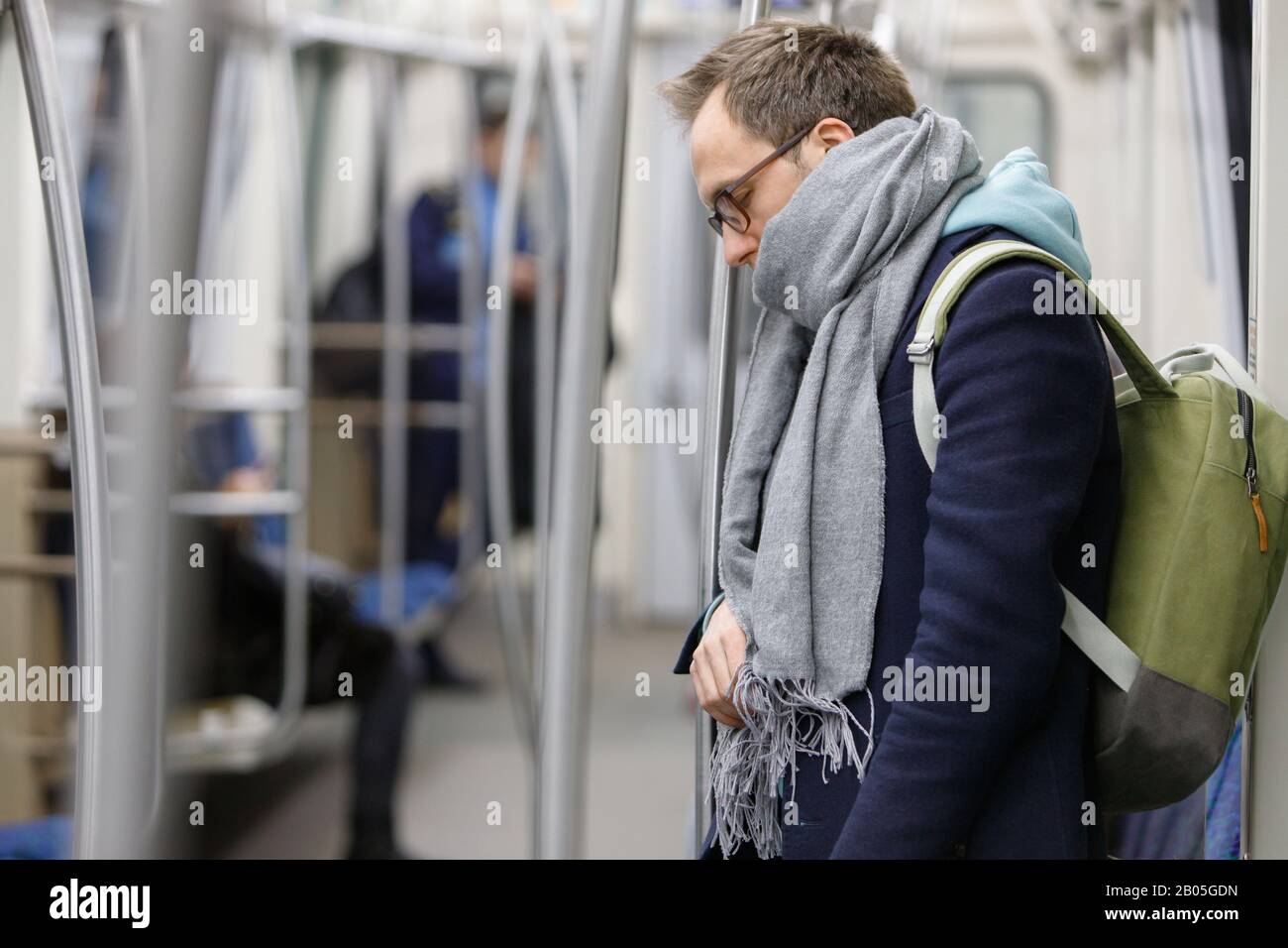 Tired man in eyeglasses in metro train. Young exhausted male standing and sleeping in subway after work, people on background. Fatigue, sleep deprivat Stock Photo