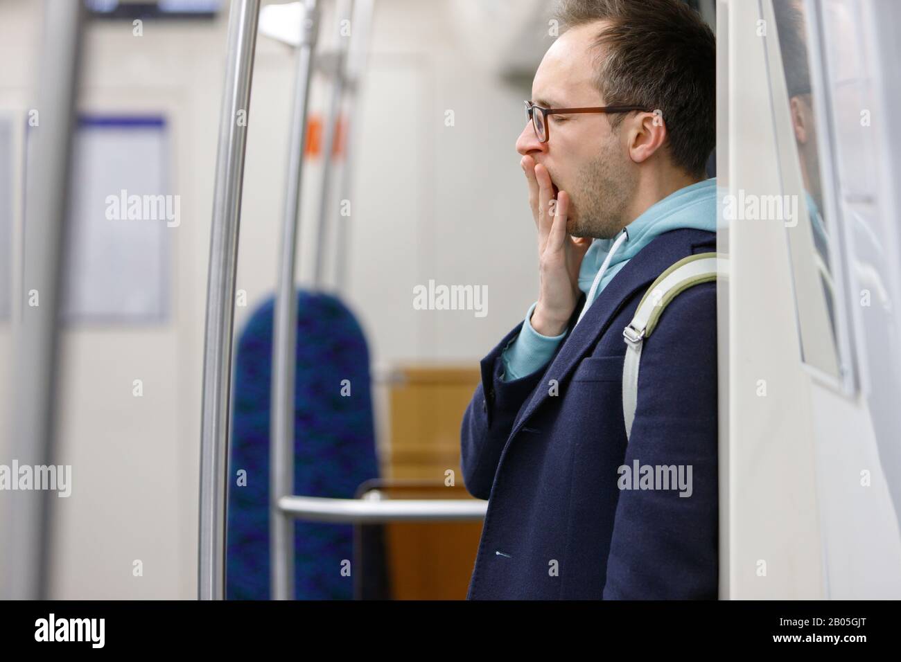 Tired man in eyeglasses in public transportation. Young exhausted male standing and yawning in subway, sleepy after work goes home, blurred background Stock Photo