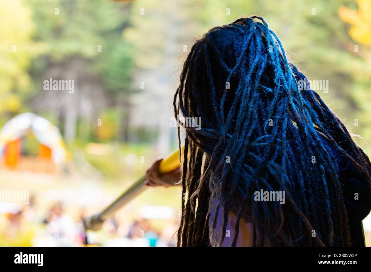 A close up selective focus shot of a woman with blue dreadlocks playing a didgeridoo. during a native culture and music festival with blurry trees in background Stock Photo