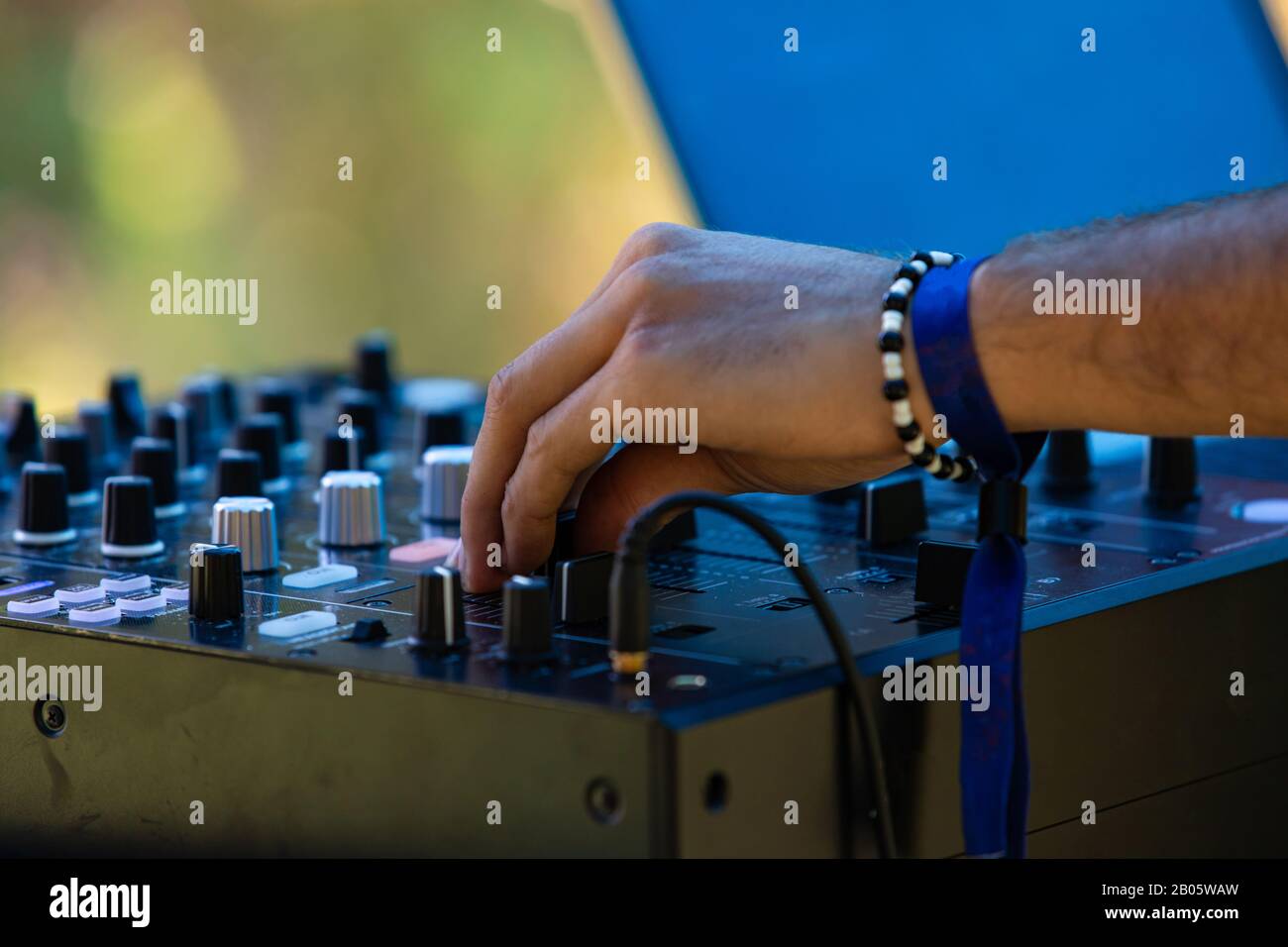 A close up view on the hand of an electronic music DJ turning dials on a CDJ mixer, wearing bangles against green blurry background at earth festival Stock Photo