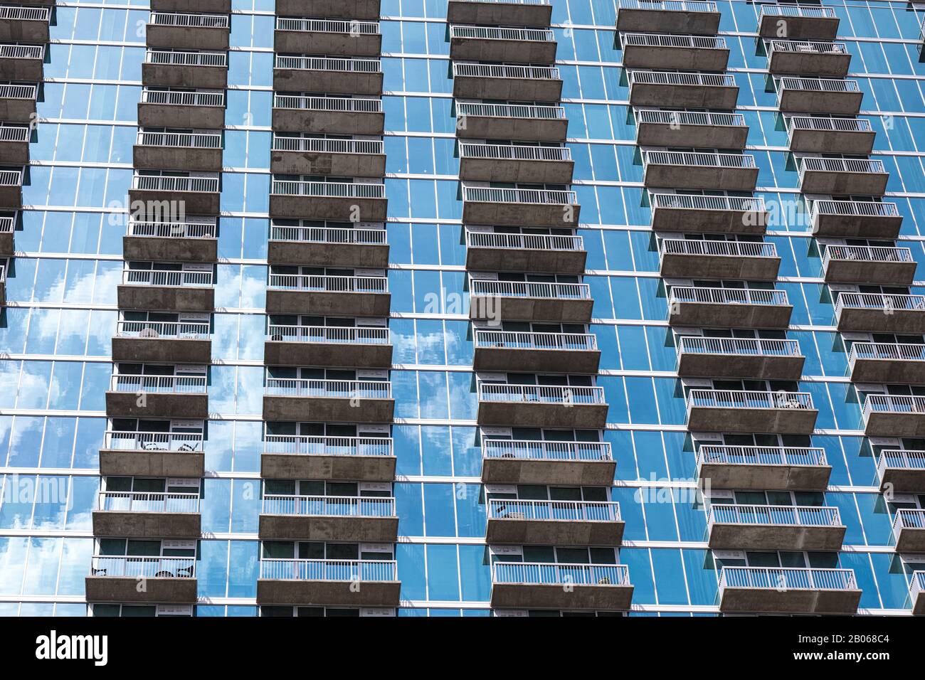 Rows of balconies forming a repetitive pattern on the facade of the modern high rise building made out of the blue glass with sky reflection on it Stock Photo