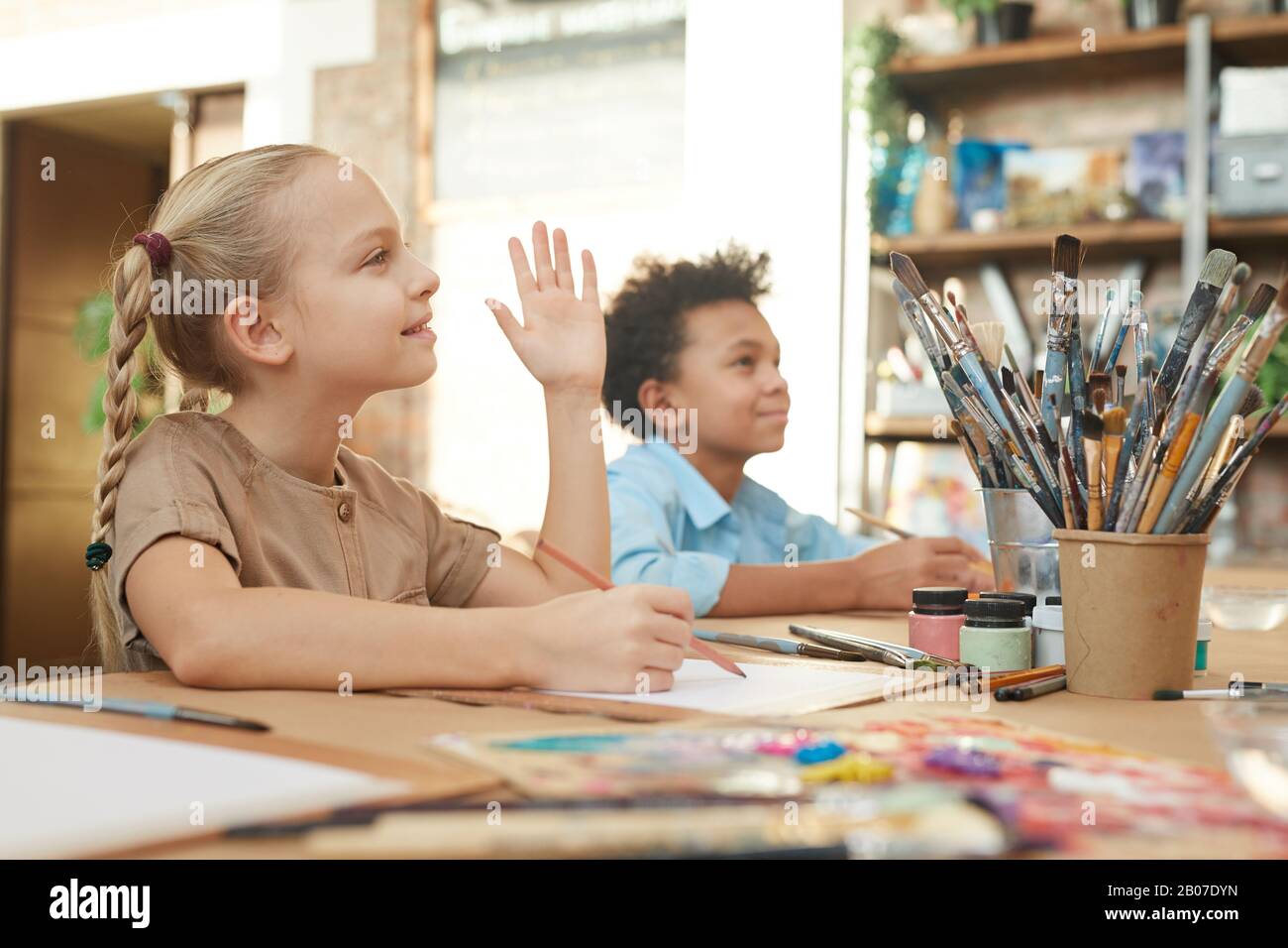 Little girl raising her hand while sitting at the table and painting ...