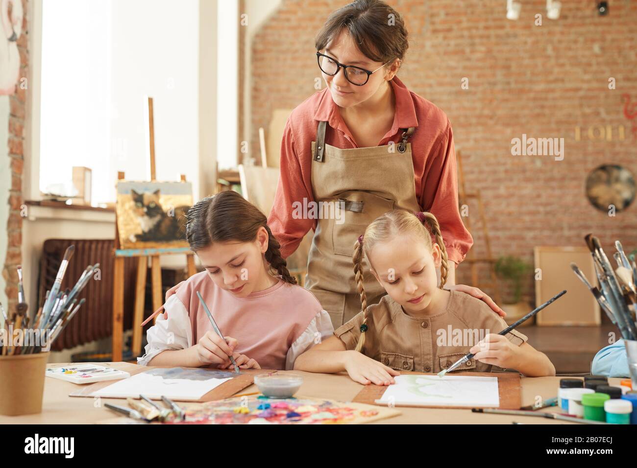 Young teacher in eyeglasses looking at work of little girls who sitting at the table and painting during art lesson at school Stock Photo