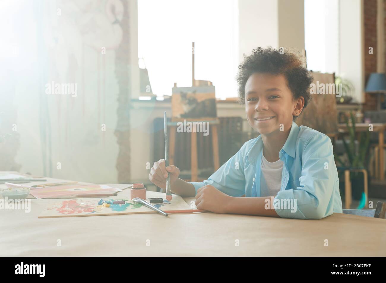 Portrait of African young boy smiling at camera while sitting at the table and drawing a picture with paints and brush Stock Photo