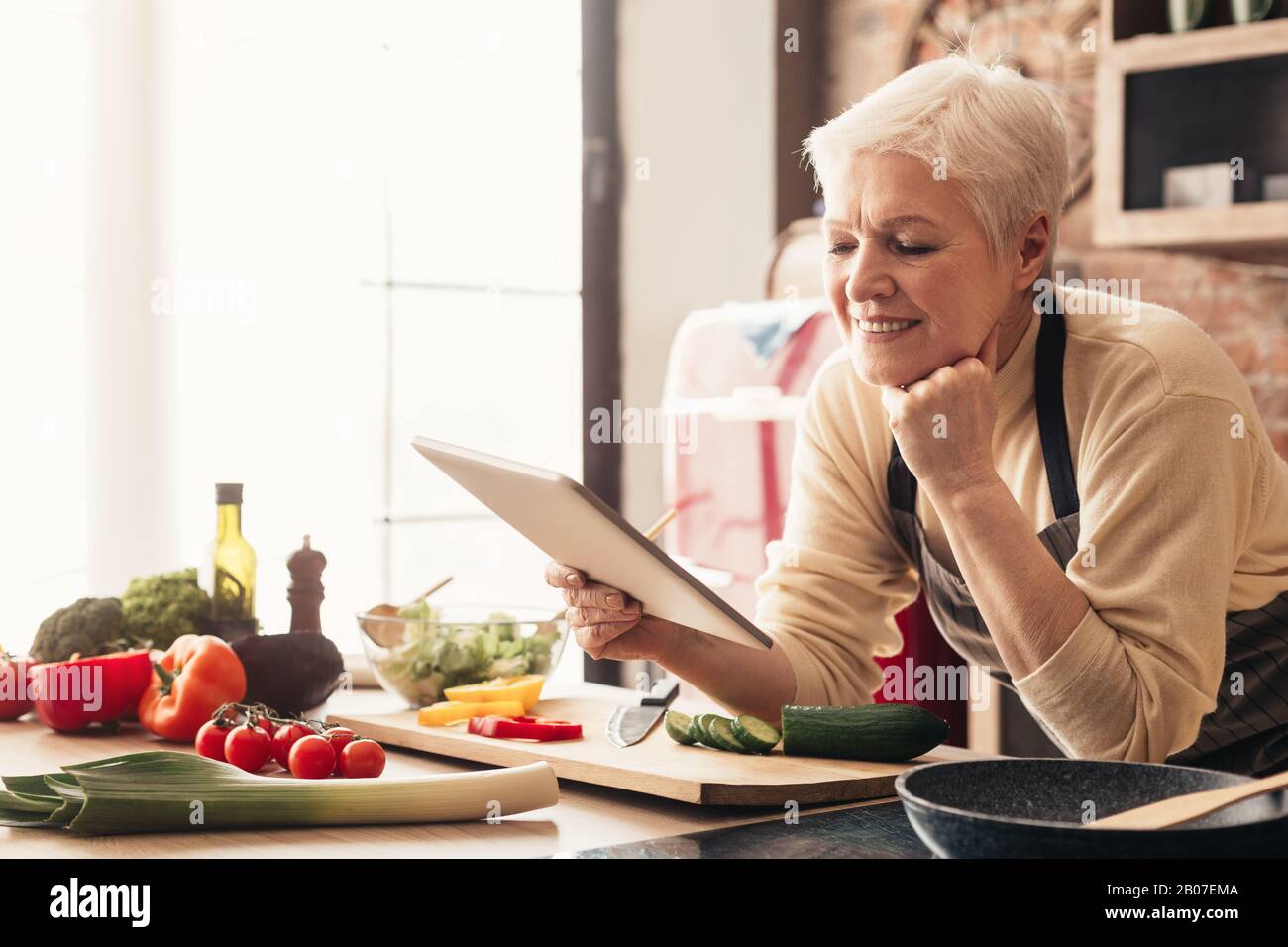 Elderly woman using digital tablet in kitchen, checking cooking recipes Stock Photo
