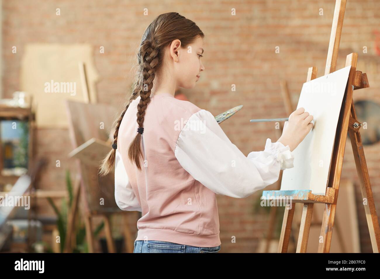 Little girl standing in front of easel and painting on it with paints ...