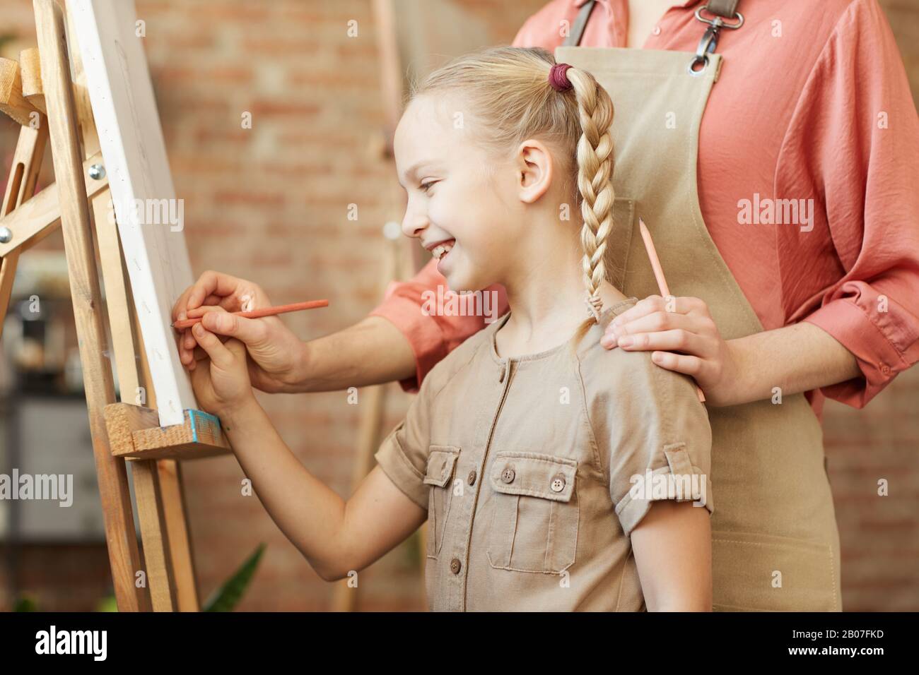 Smiling little girl standing in front of easel and learning to paint ...