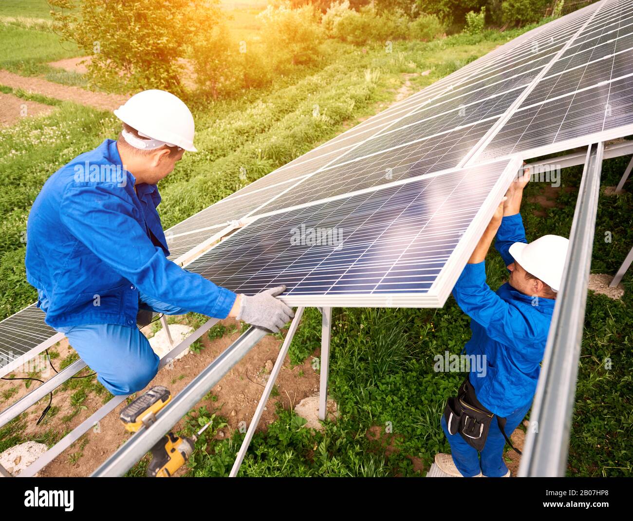 Installing of stand-alone solar photo voltaic panel system. Two technicians in hard-hats mounting big shiny solar module on platform on green summer view background. Alternative energy concept. Stock Photo