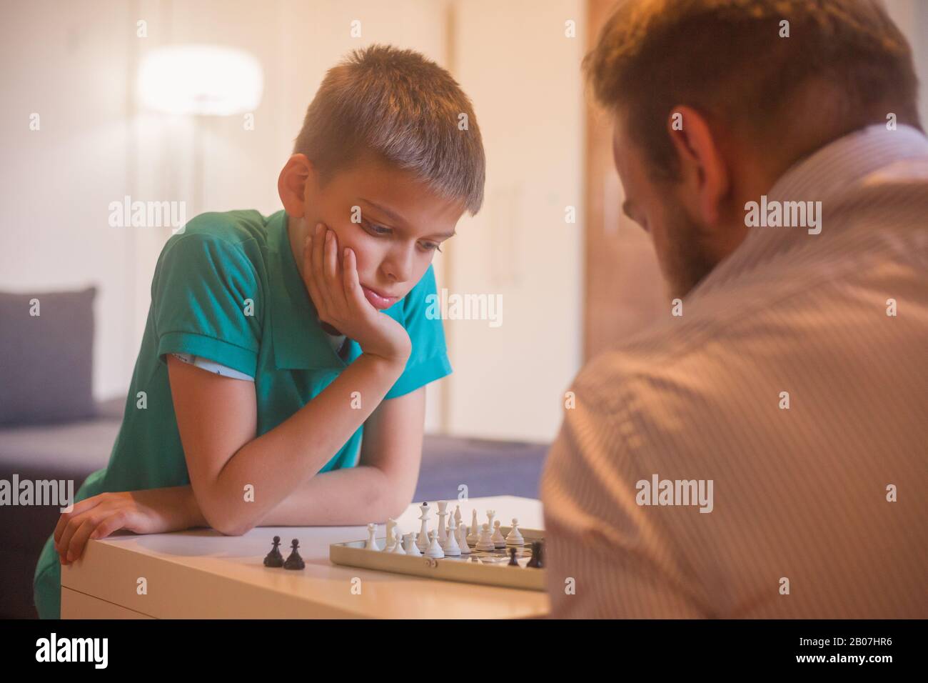 young man and son playing chess together. Son is thinging about his next move. Stock Photo