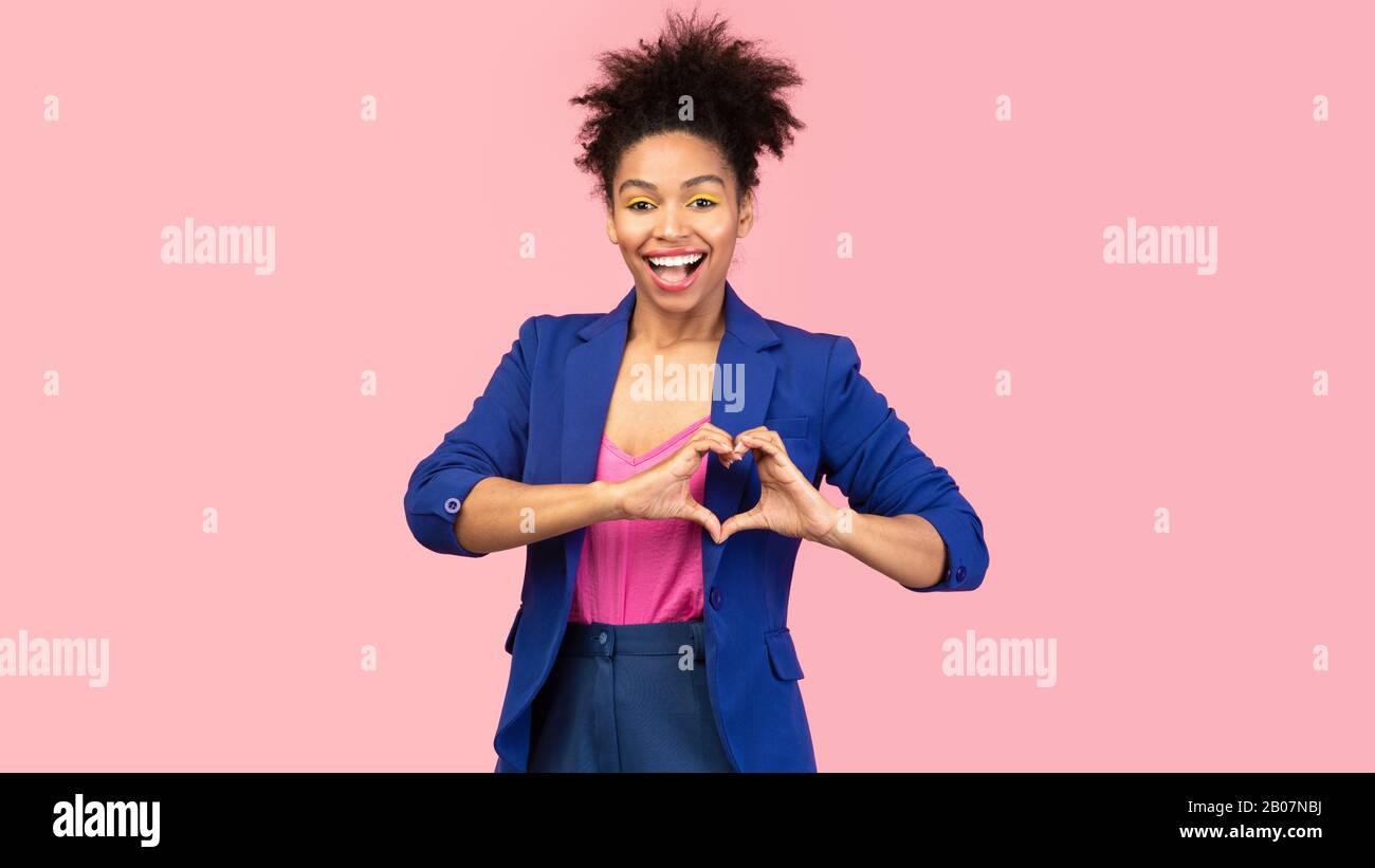 Smiling afro girl showing heart gesture at pink studio Stock Photo