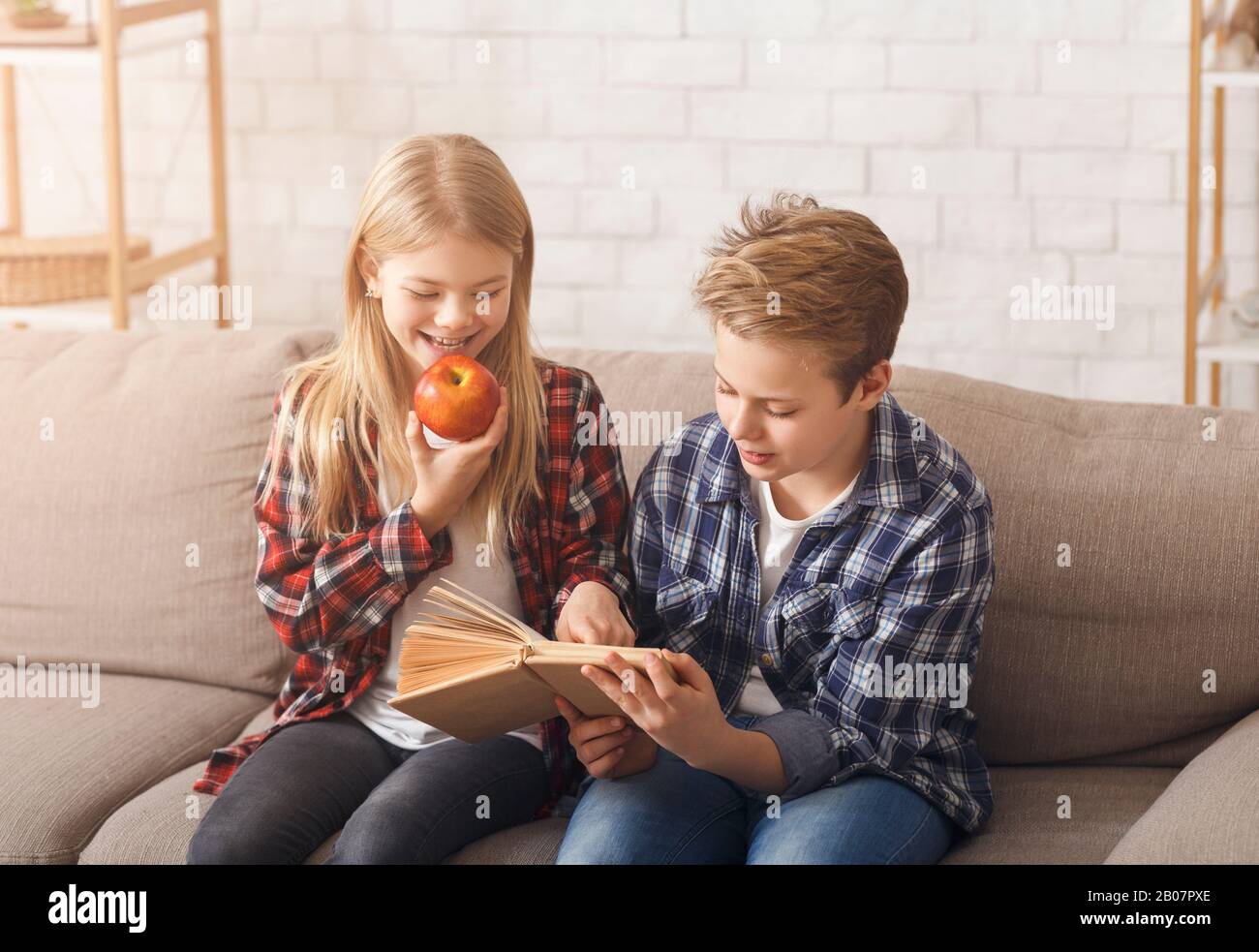 Brother And Sister Reading Book Sitting On Sofa At Home Stock Photo