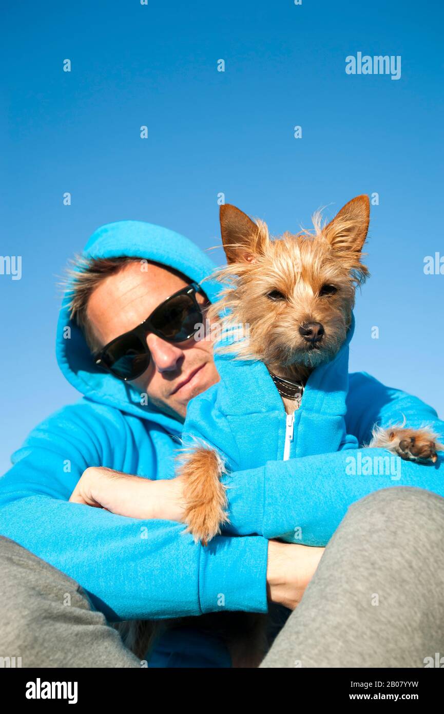 Man sitting with dog in matching blue hooded sweatshirts under bright sunny sky Stock Photo