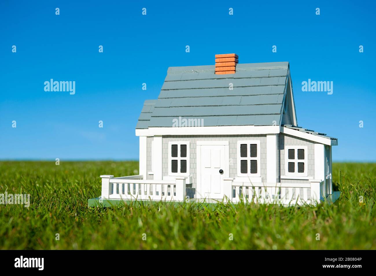 Miniature house with white picket fence standing in lush green grass lawn in front of a blue sky horizon Stock Photo