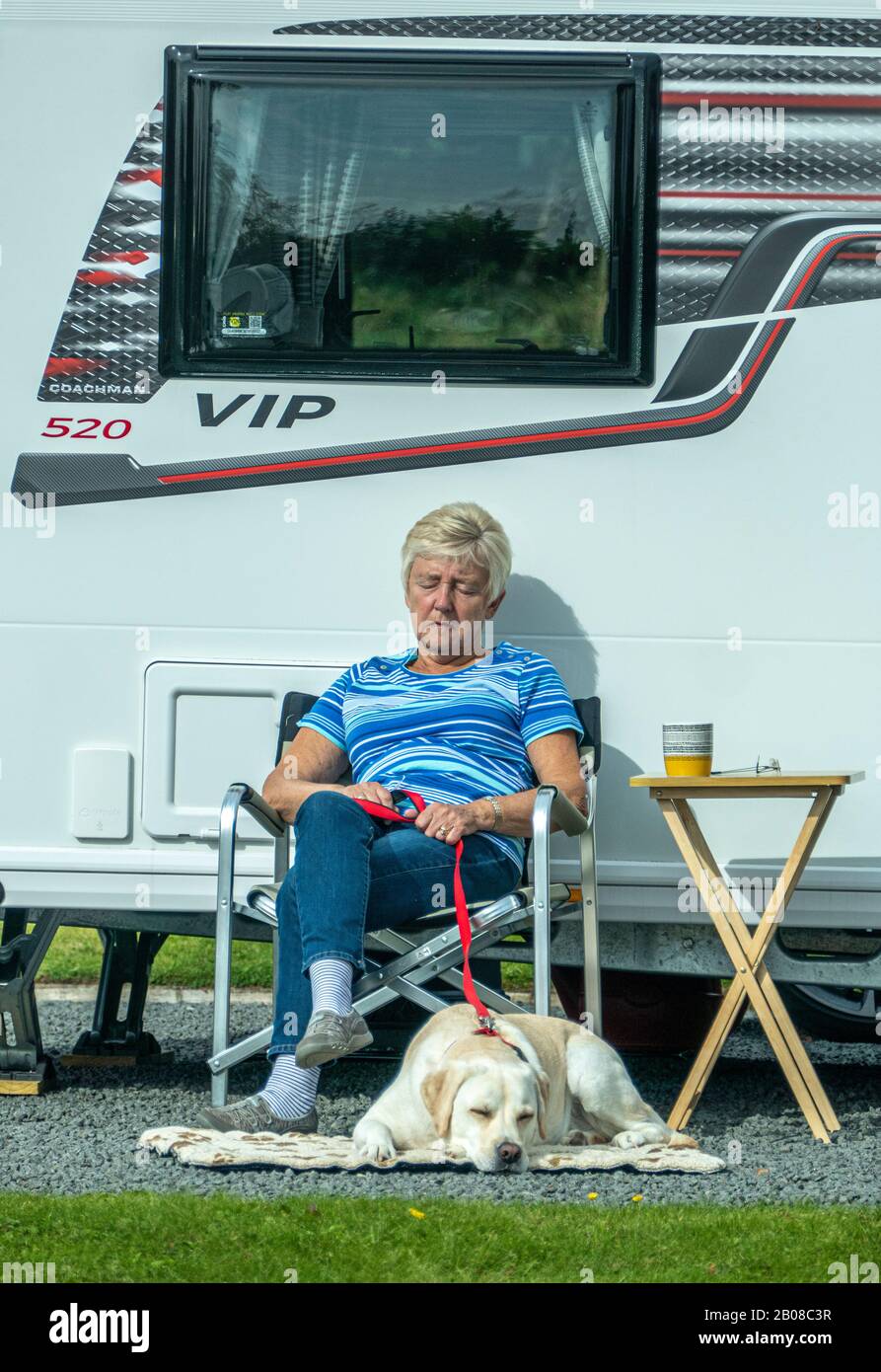 Elderly / senior woman sitting in a chair in bright sunshine outside her caravan, on a peaceful campsite, with her pet labrador dog lying next to her. Stock Photo
