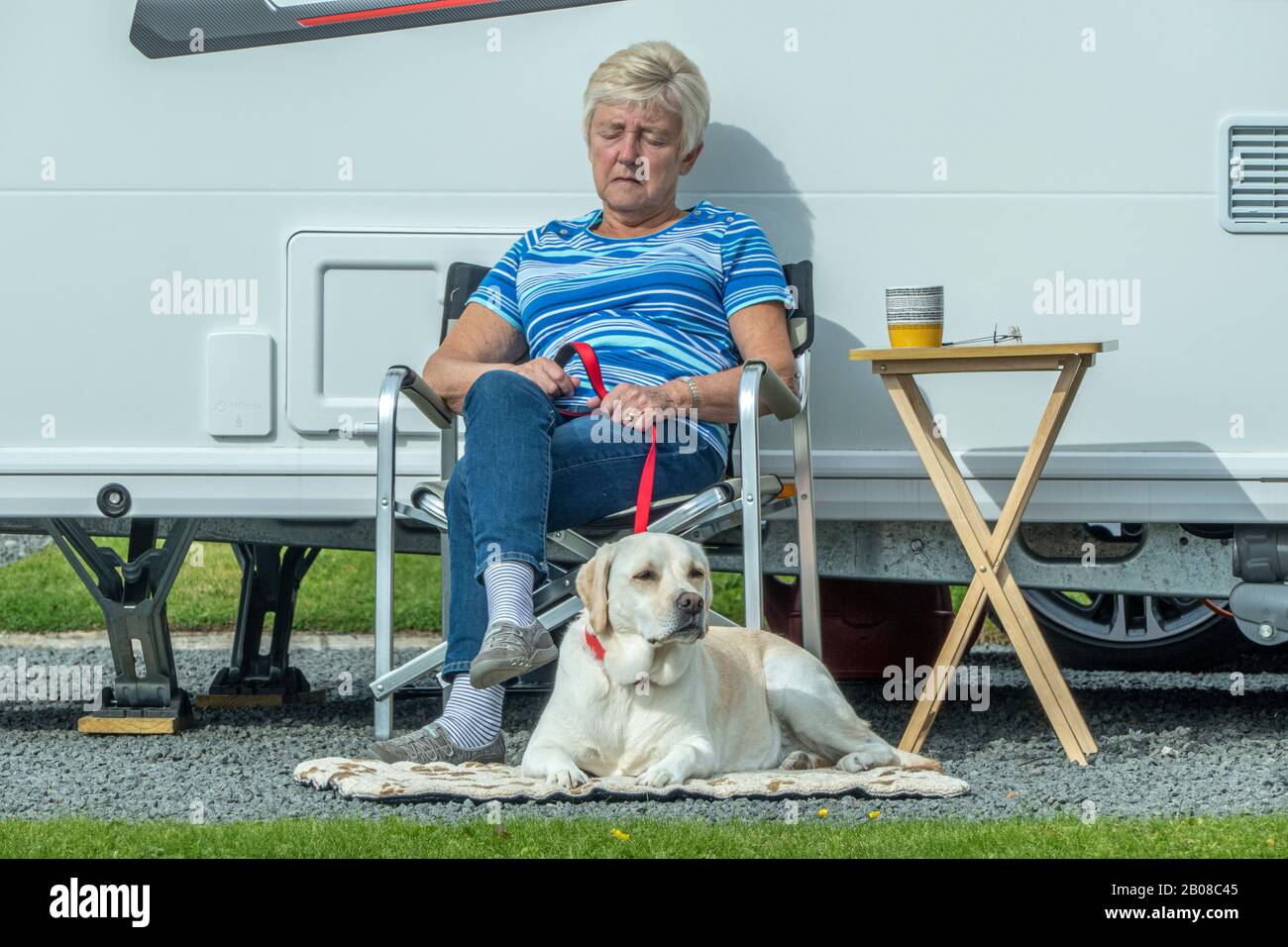 Elderly / senior woman sitting in a chair in bright sunshine outside her caravan, on a peaceful campsite, with her pet labrador dog lying next to her. Stock Photo