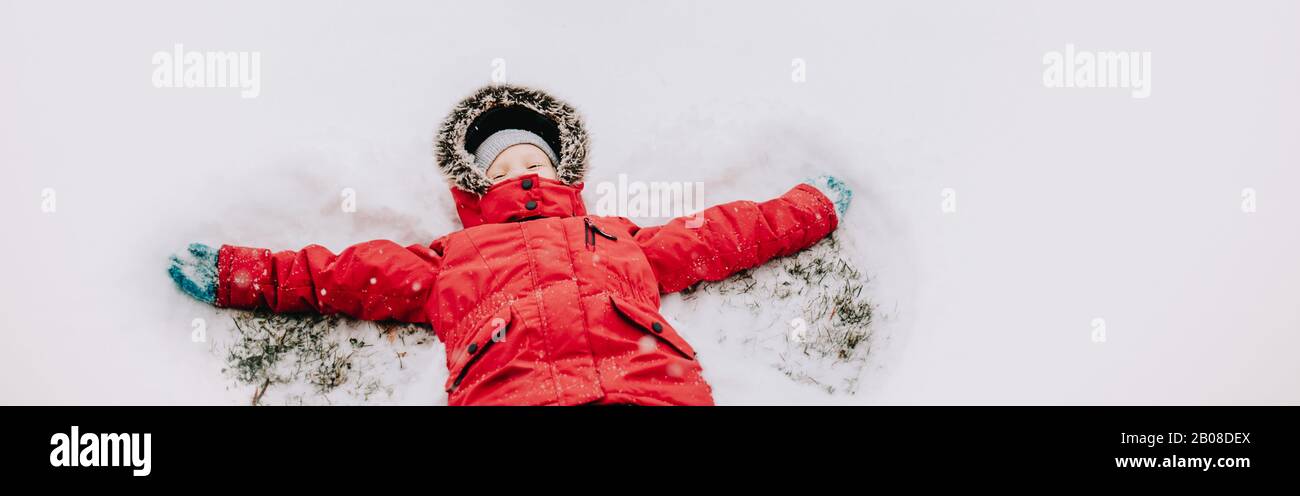 Cute adorable Caucasian girl child in warm clothes red jacket lying on snow ground during cold winter snowy day. Kids outdoor seasonal activity. Child Stock Photo