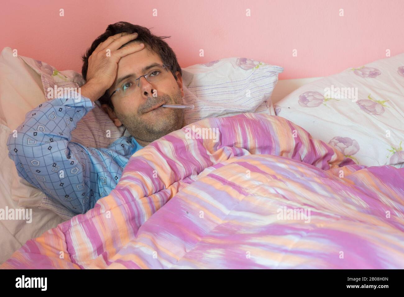 Portrait of a sick young man lying in bed taking temperature and having flu. Stock Photo