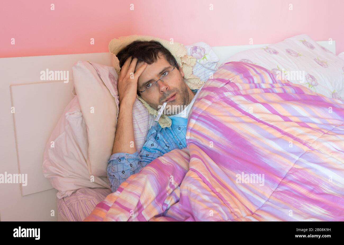 Portrait of a sick young man lying in bed taking temperature and having flu. Stock Photo