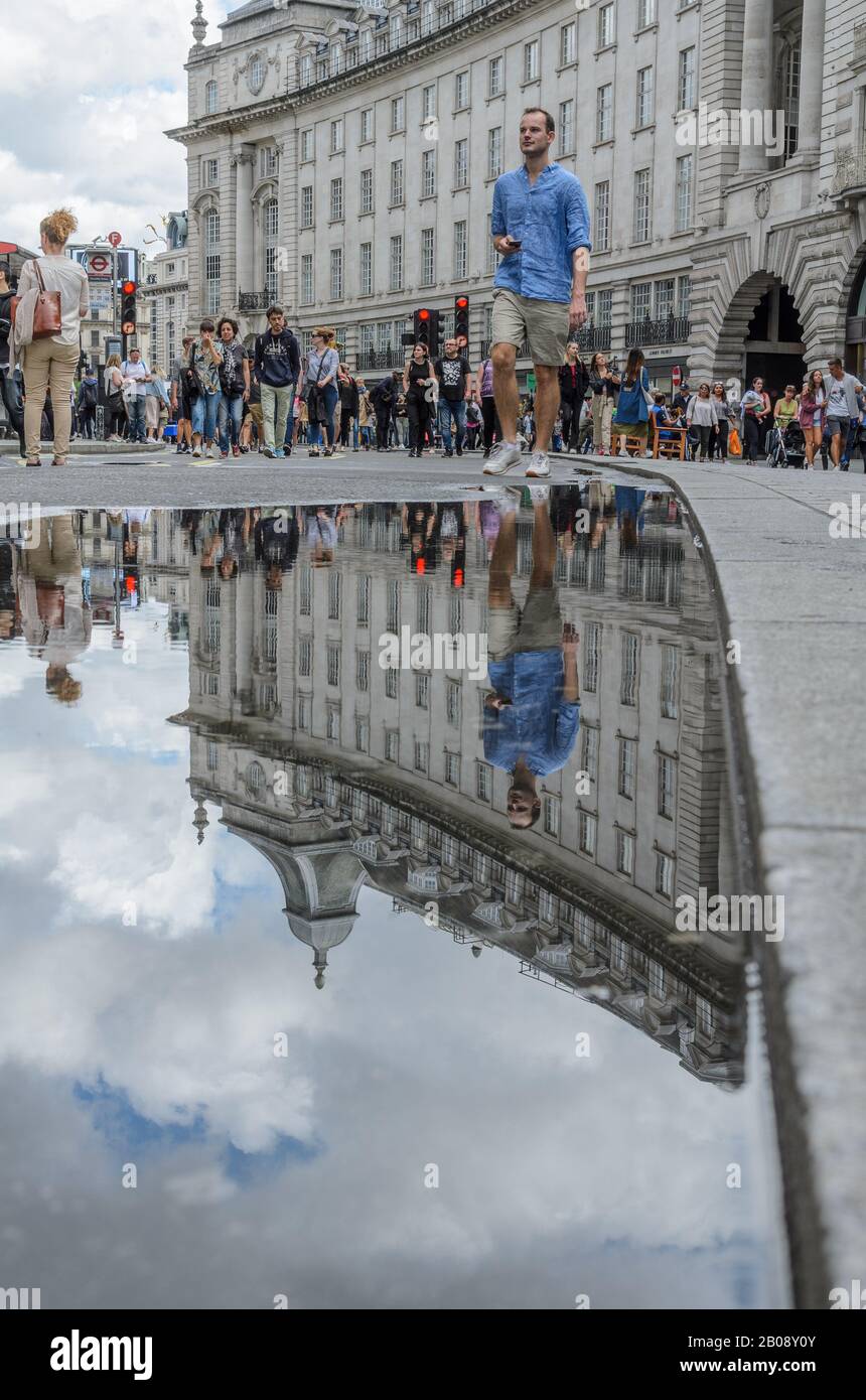 A man in a blue shirt and shorts reflected in a puddle on a busy summer shopping day on Regent Street in London Stock Photo