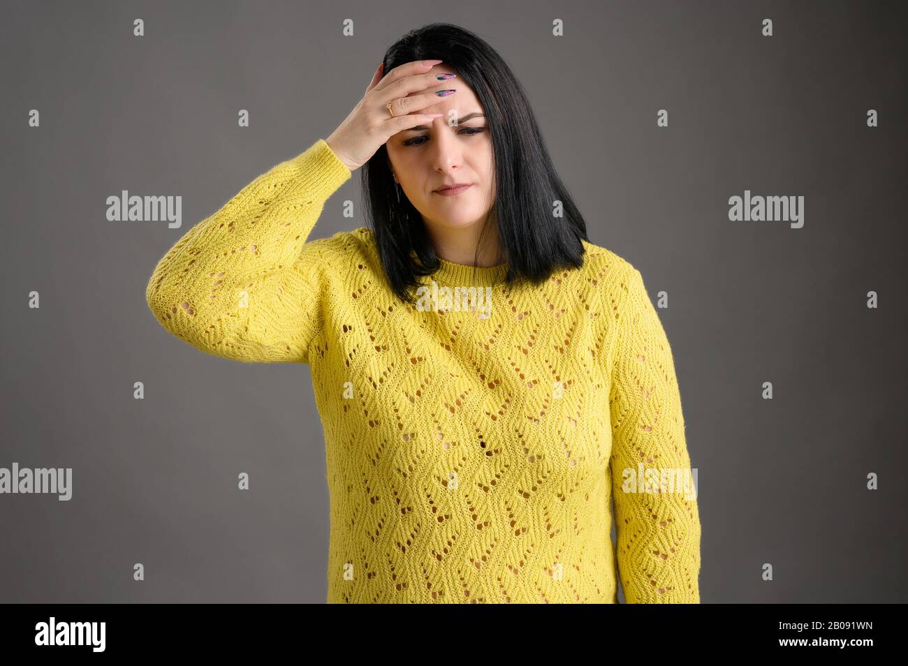 Young woman with black hair dressed casually in a yellow sweater has headaches posing an isolated grey background Stock Photo