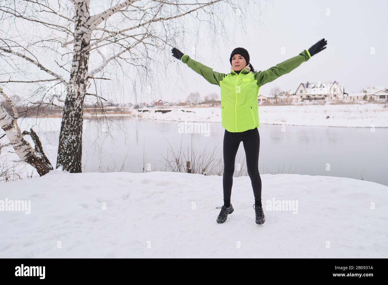 Active young woman in sportswear doing jumping exercise with raised arms at winter lake Stock Photo