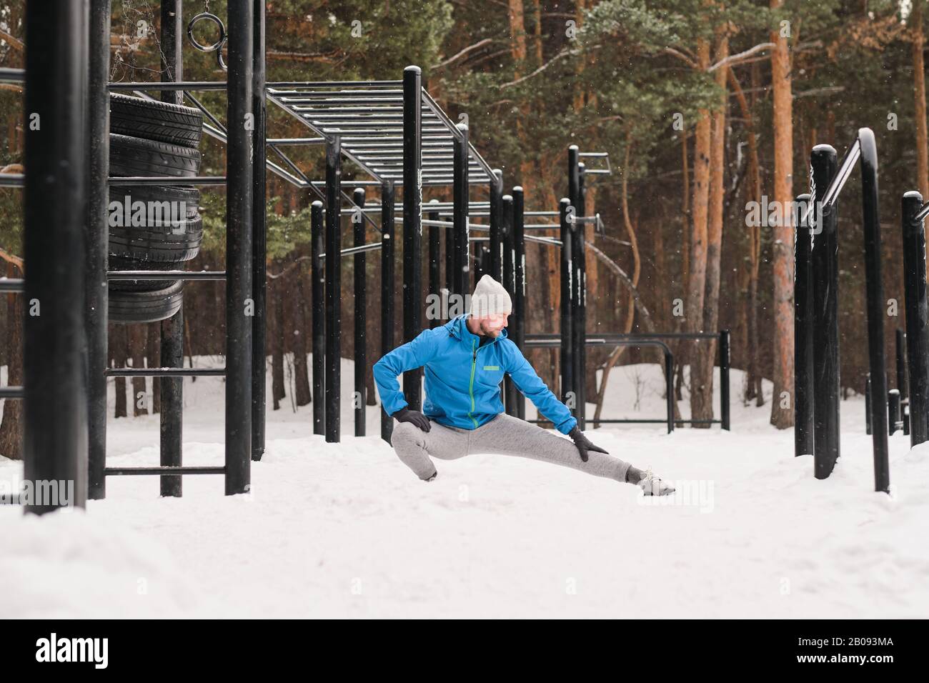 Young man in warm clothes doing crouch adductor stretch while exercising at winter sports ground Stock Photo