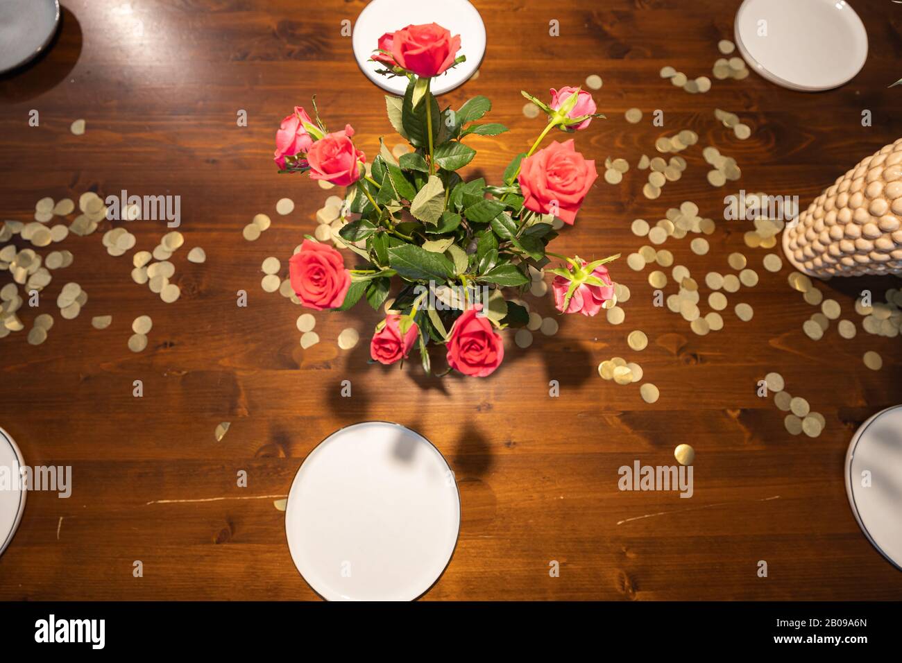 Top down view of a Vase with red roses on a table with gold confetti and plates for dinner. Stock Photo