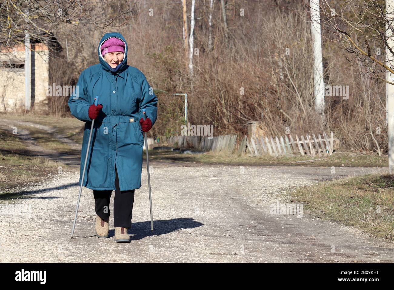Elderly woman walking with sticks on rural road, sports exercises for healthy spine and joints. Nordic walking at cold weather, therapy for health Stock Photo