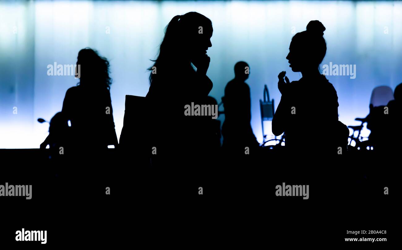 Silhouettes of two teen girls standing and talking in front of modern building, in the night, and people passing by, in high contrast black and blue Stock Photo