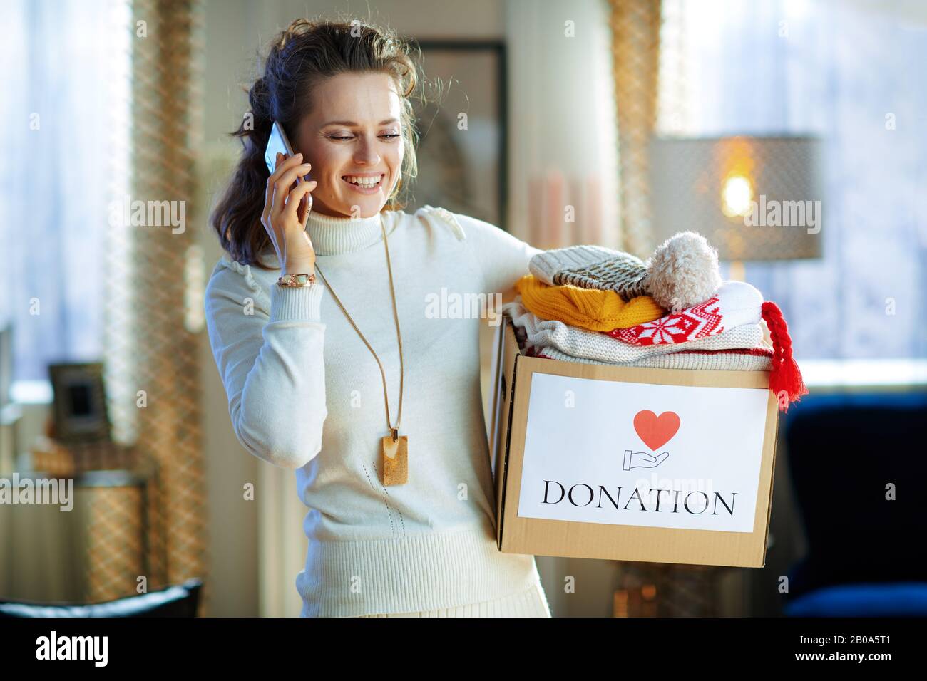 happy stylish woman in white sweater and skirt with donation box with old warm clothes talking on a cell phone at modern home in sunny winter day. Stock Photo