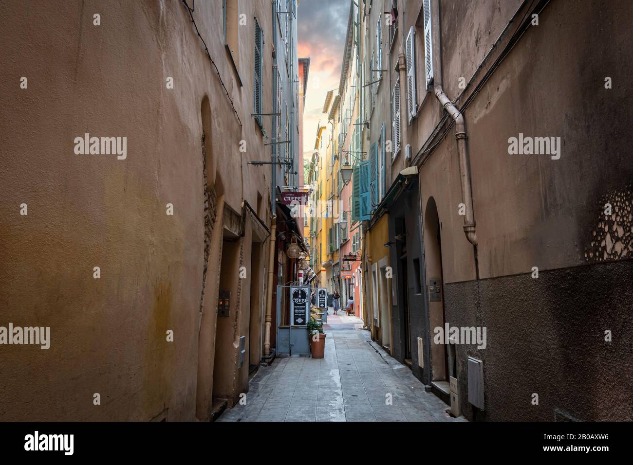 Colorful storm clouds and high walls border a narrow alley with cafes and shops in the Old Town Vieux Nice section of Nice, France. Stock Photo