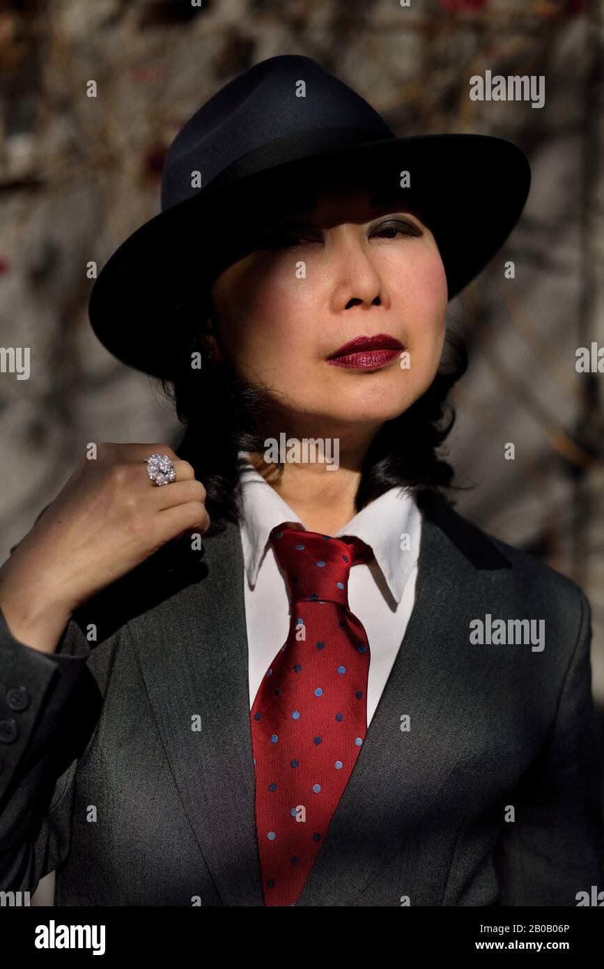 Face in partial shadow, an Asian woman wearing a mans style sharkskin suite, red necktie and fedora hat looking like a 1940s gangster, red lips Stock Photo