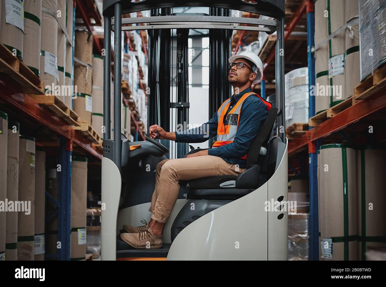 Happy forklift driver focused on carefully transporting stock from shelves around the floor of a large warehouse wearing a white helmet and vest Stock Photo