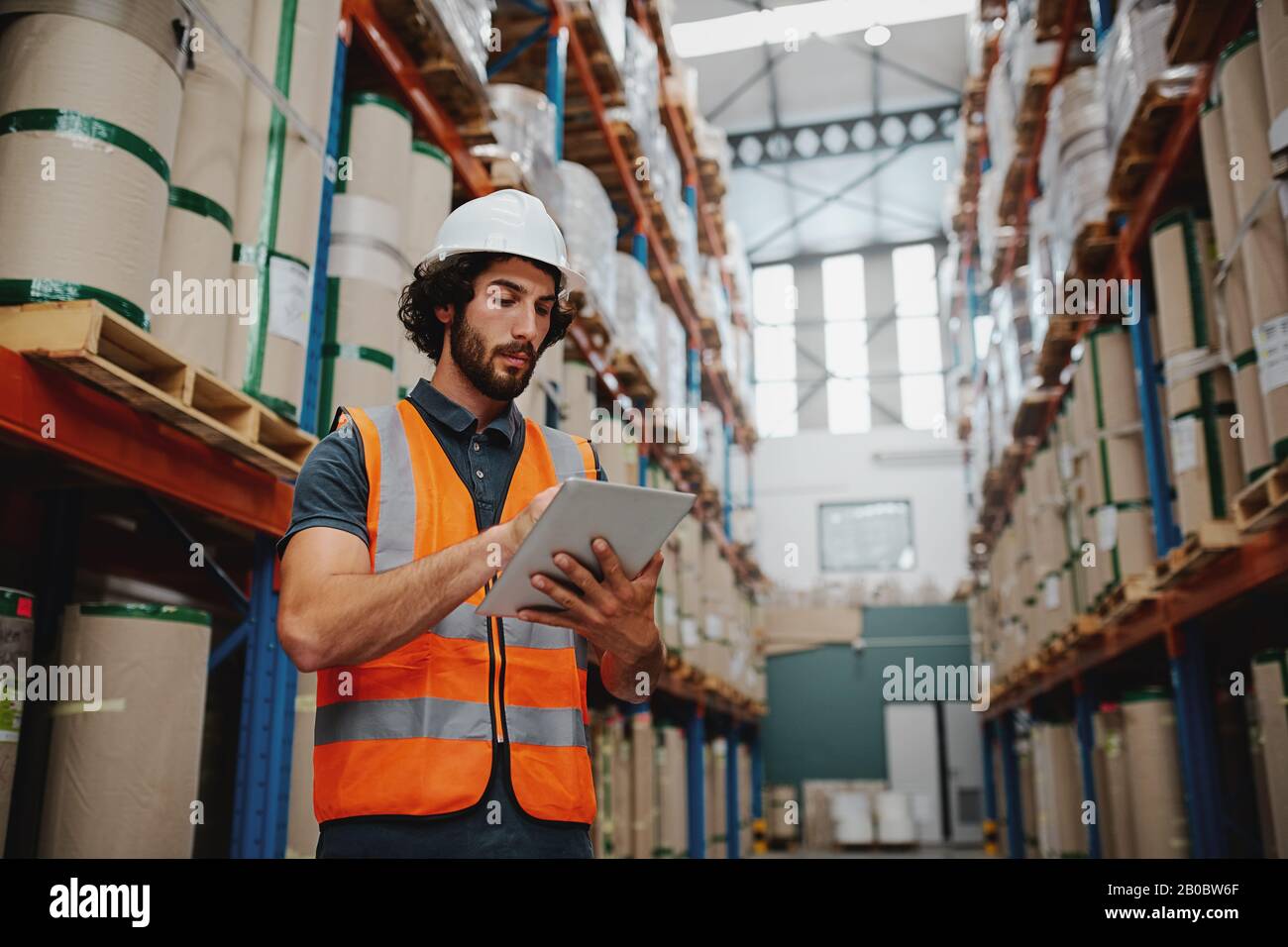 Young caucasian engineer in hardhat is using a tablet computer in a heavy industry factory wearing white helmet and orange vest for safety Stock Photo