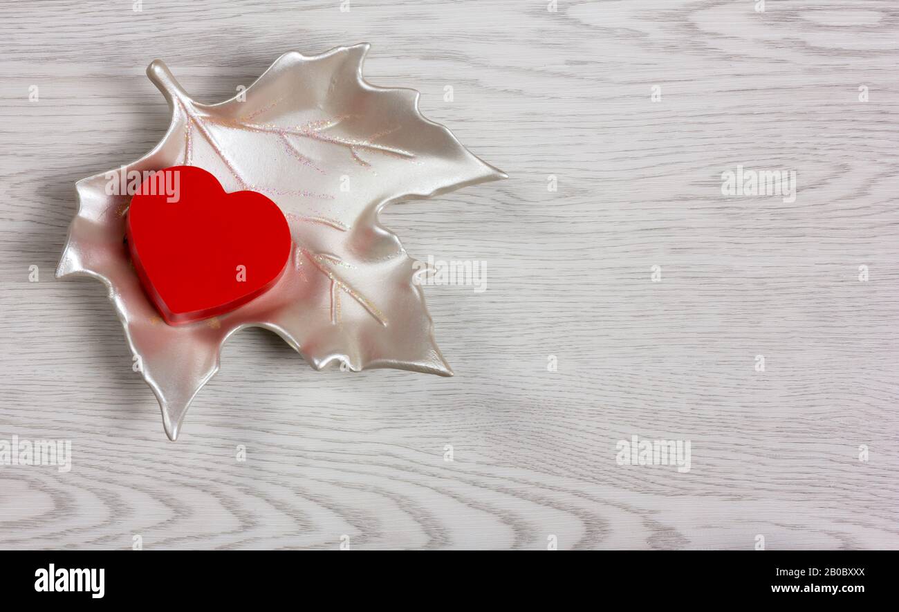 Red heart-shaped small box on a ceramic white maple leaf over a white wooden background Stock Photo