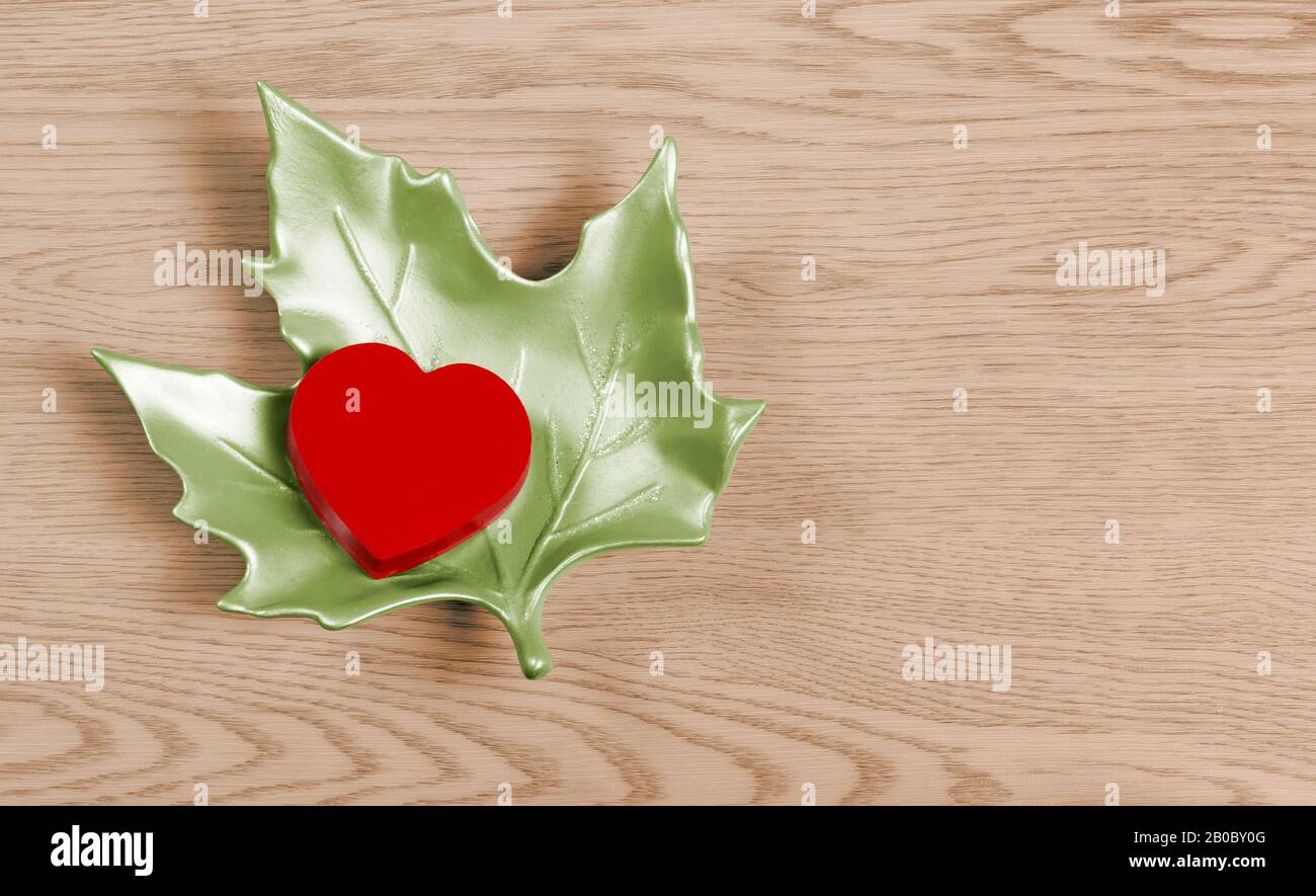 Red heart-shaped small box on a ceramic green maple leaf over a wooden background Stock Photo