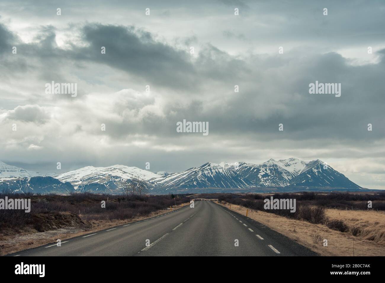 Empty road leads toward a snow capped mountain with moody, cloud sky. Stock Photo