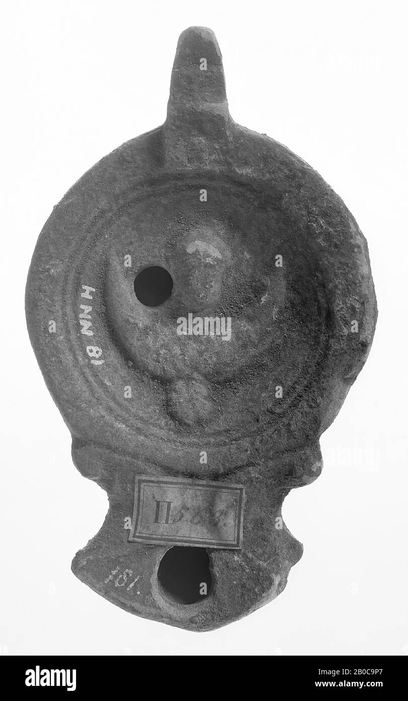 A black-nested oil lamp with a round body on a recessed, round base. The concave mirror is surrounded by recessed concentric circles and is decorated with a winged bust with the small filling hole to the left of it. The short, broad spout with a small fire hole is angularly closed and framed with volutes. The ear is turned on vertically., Oil lamp, pottery, terracotta, 3 x 10.4 x 6 cm, 1st and 2nd century AD. 1-200 AD, Tunisia Stock Photo