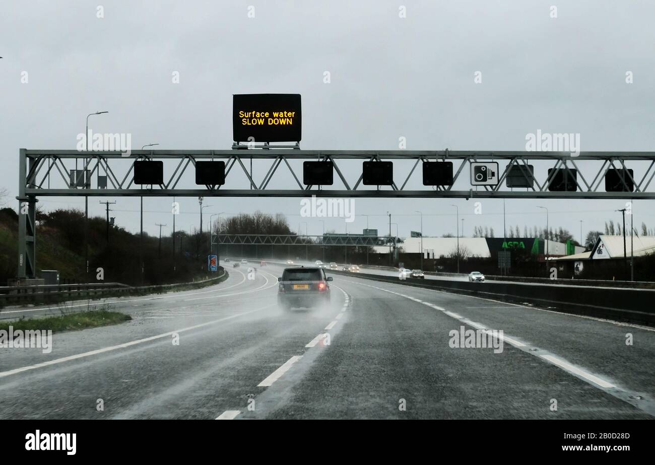 A car drives along a UK motorway, in heavy rain, with a sign board on a gantry above the motorway advising motorists to slow down for surface water. Stock Photo