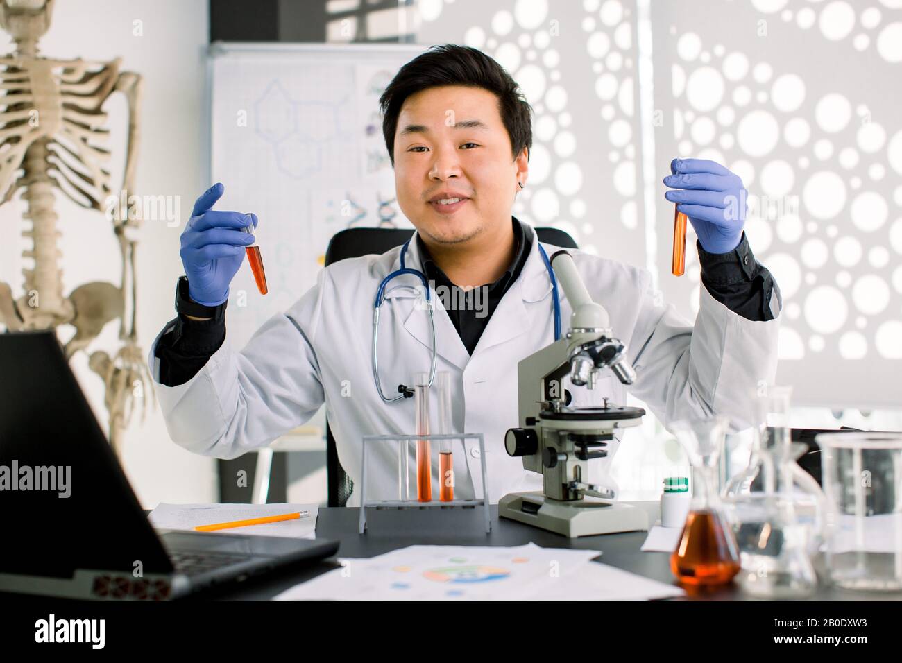 Professional smiling Asian scientist doctor, laboratory man, holding two test tubes with blood samples, while making a research and using microscope Stock Photo