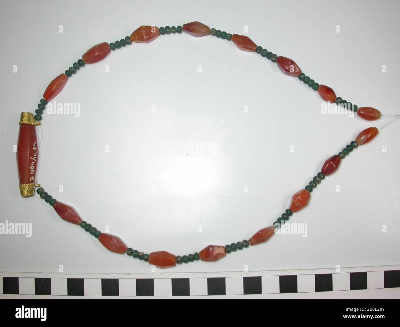 Necklace of round, elliptical and polygonal beads of glass and stone. Two elliptical beads of carnelian stone, 14 polygonal beads of carnelian stone and 64 small round beads of green glass, always with four between the stone beads. Finally, at the bottom of the middle, a very long polygonal bead of carnelian stone between cap-like golden catches with pearl edges, rosettes and suspension eyes. All beads completely and almost flawlessly preserved, everything on a modern cord strung. Total: 81 beads., Ornament, glass, metal, gold, stone, L 24 cm, 600-200 BC, Iran Stock Photo