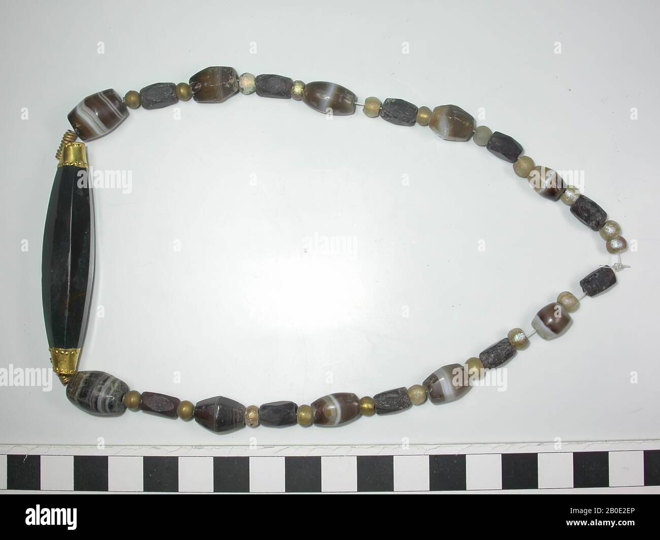 Necklace of round, barrel-shaped, polygonal and yet other beads of glass, stone and gold. Fully symmetrical of construction. 16 small round beads of colorless, but gold-yellow iridescent glass, 10 polygonal beads of dull black stone, 8 barrel-shaped beads of brown and white sardonyx like layered stone, 2 polygonal beads of the same kind of stone and 4 small round golden beads with profiling on top and bottom. Finally, after 2 elongated spiral golden beads, a very long polygonal bead of dark green stone in a middle setting with pressed pearl edges, rosettes and suspension eyes. All beads are Stock Photo