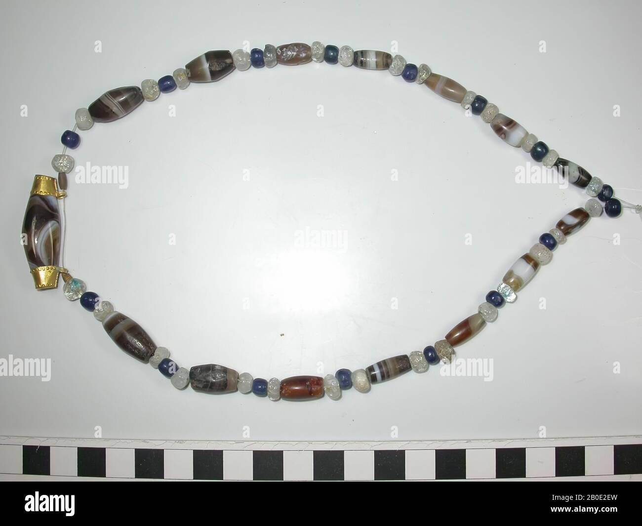 Necklace from round to flattened round and also long, cylindrical beads of stone, glass and gold. 16 small round beads of blue glass, 30 small round to flat beads of colorless to milky white glass or stone, 14 downwards cylindrical beads of brown and white sardonyx like layered stone, 2 small elongated beads of gold and finally at the center bottom one very long cylindrical bead of brown and white sardonyx-like layered stone, between two golden rings with pearls and rosettes and eyes. All beads are almost perfectly preserved. Well strung on modern cord. Total: 63 beads, ornament, metal, gold Stock Photo