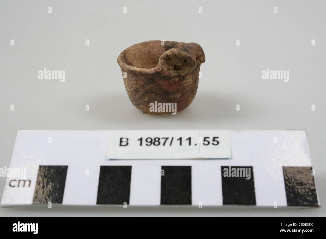 An earthenware miniature bowl with flat bottom. The wall rises steeply and bulges out slightly. The edge is bent outwards and unthickened. On the edge there is a small modeled animal head with a rounded down horn on either side. The ears are indicated by a round hole. The outside has a beige-green coating with a dark brown paint. A double horizontal line is placed under the edge and three horizontal zigzag lines on the belly. On the top and horns of the animal head we see cross stripes. The interior has a beige coating, crockery, earthenware, handmade, H 3.3 cm, D 3.3 cm, Bronze Age 1800-1150 Stock Photo