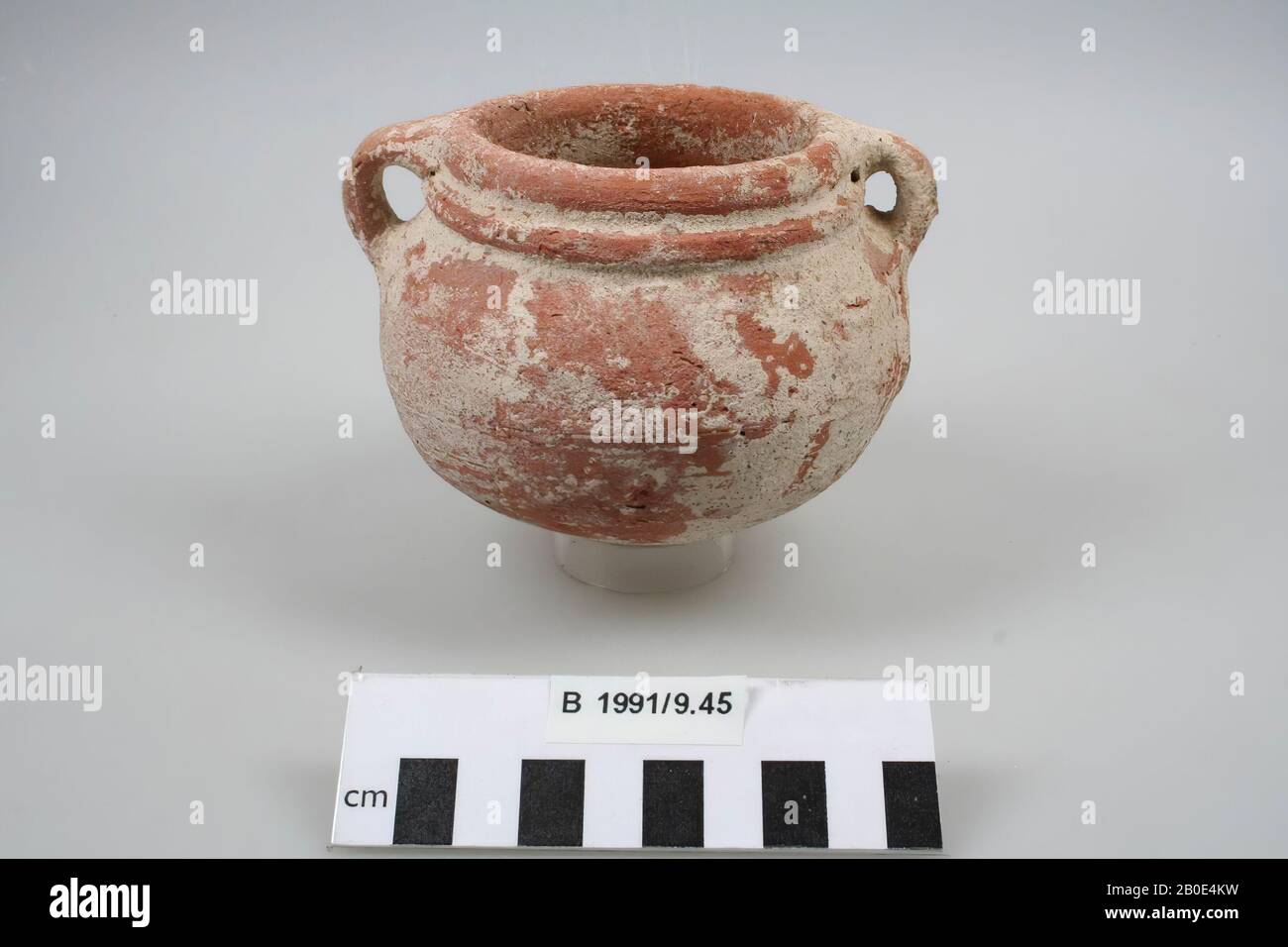 Small miniature cooking pot of baked clay with a round bottom that runs downwards. The body is bulging. The neck is low and wide. The edge is thickened on the outside. On the neck is a large horizontal ledge. Two vertical handles are attached to the rim and the shoulders. Manufactured on the wheel. The clay is red-orange in color, has a fine texture and is agglutinated with lime and medium-coarse sand. On the outside a red-orange coating of which large parts are covered with white deposits. The surface contains horizontal spinning traces, prints of, inter alia, plant inclusions and some small Stock Photo