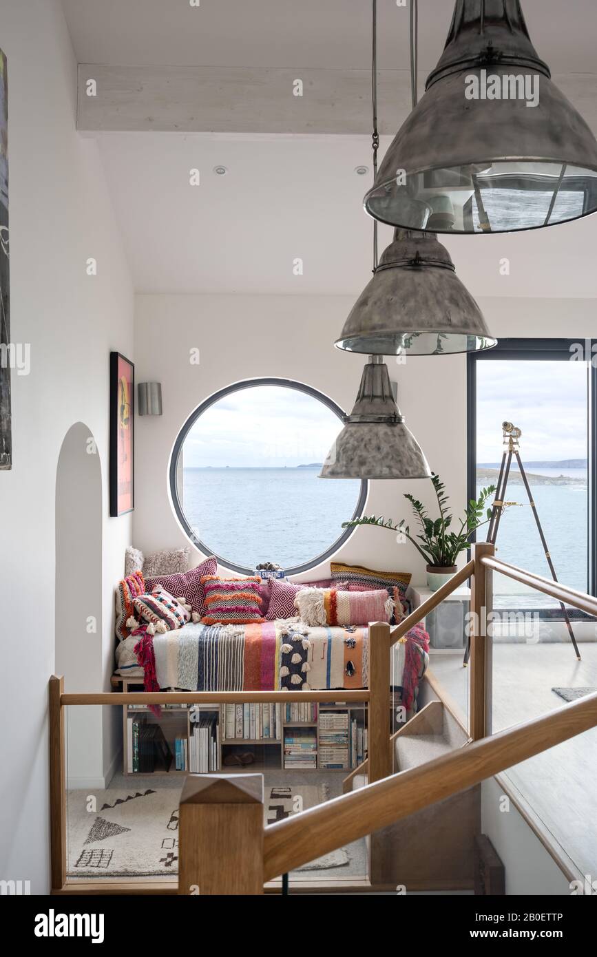 Large porthole window above daybed and storage bench in split level 1960s renovation with view to Fistral Beach Stock Photo